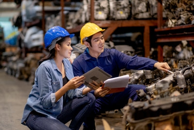 Two engineers in safety helmets discussing parts in an automotive warehouse, focused on teamwork and inventory management.